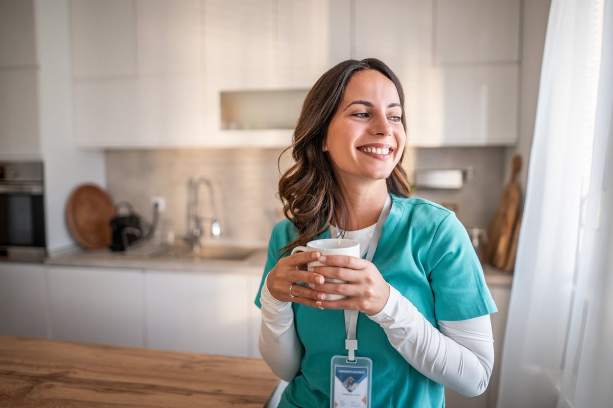 Female Continence Nurse Specialist in scrubs, smiling with coffee in a modern kitchen